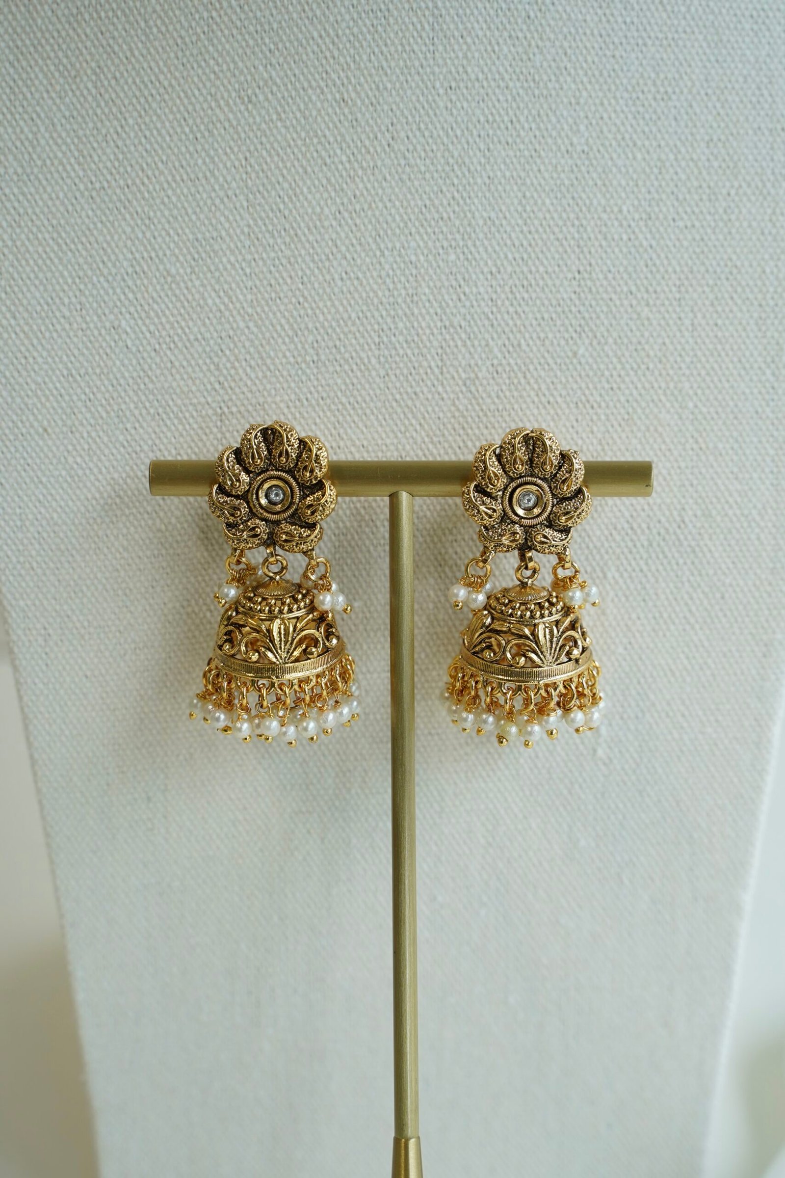 Pair of matching antique matte gold jhumka earrings featuring an intricate floral top design and tiny hanging pearl beads.