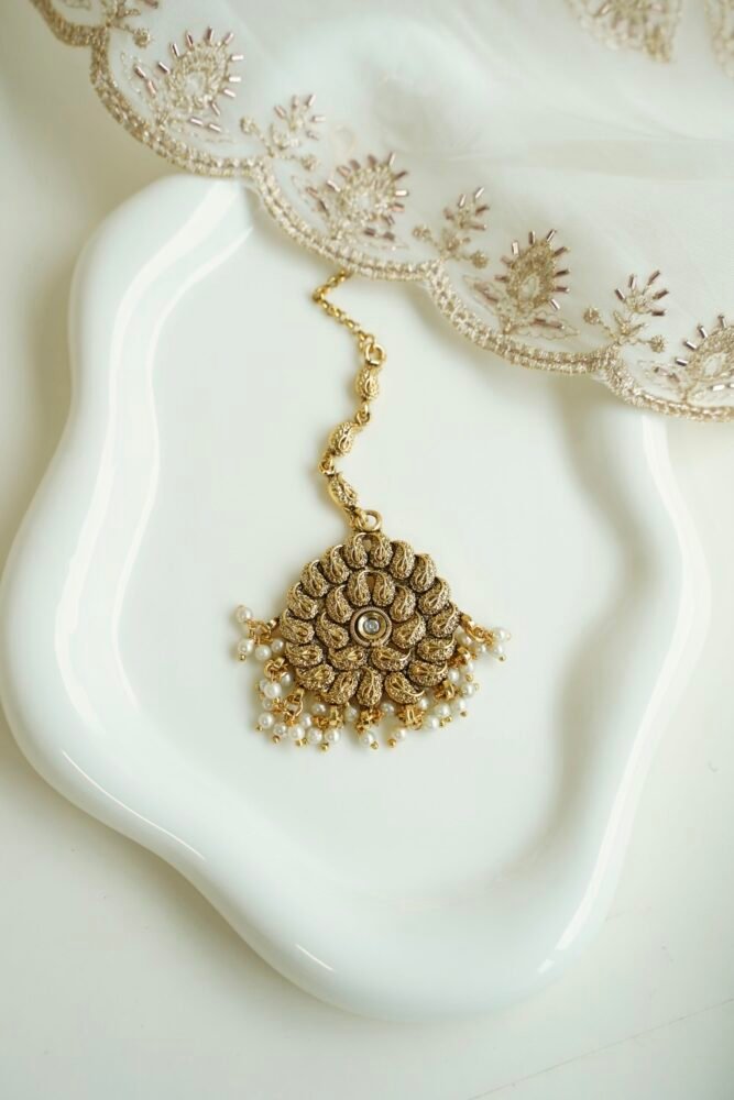 Detailed close-up of a circular antique gold maang tikka with a central crystal stone and hanging delicate white pearl clusters.