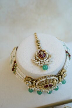 Aaliyah choker set in maroon and green with multi-strand pearl beaded band, antique gold centerpiece, mint green bead drops, jhumka earrings and tikka displayed on a bust